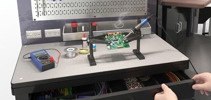 La misma mesada montada para electrónica: una placa sujeta en un soporte, un soldador, estaño y un multímetro.