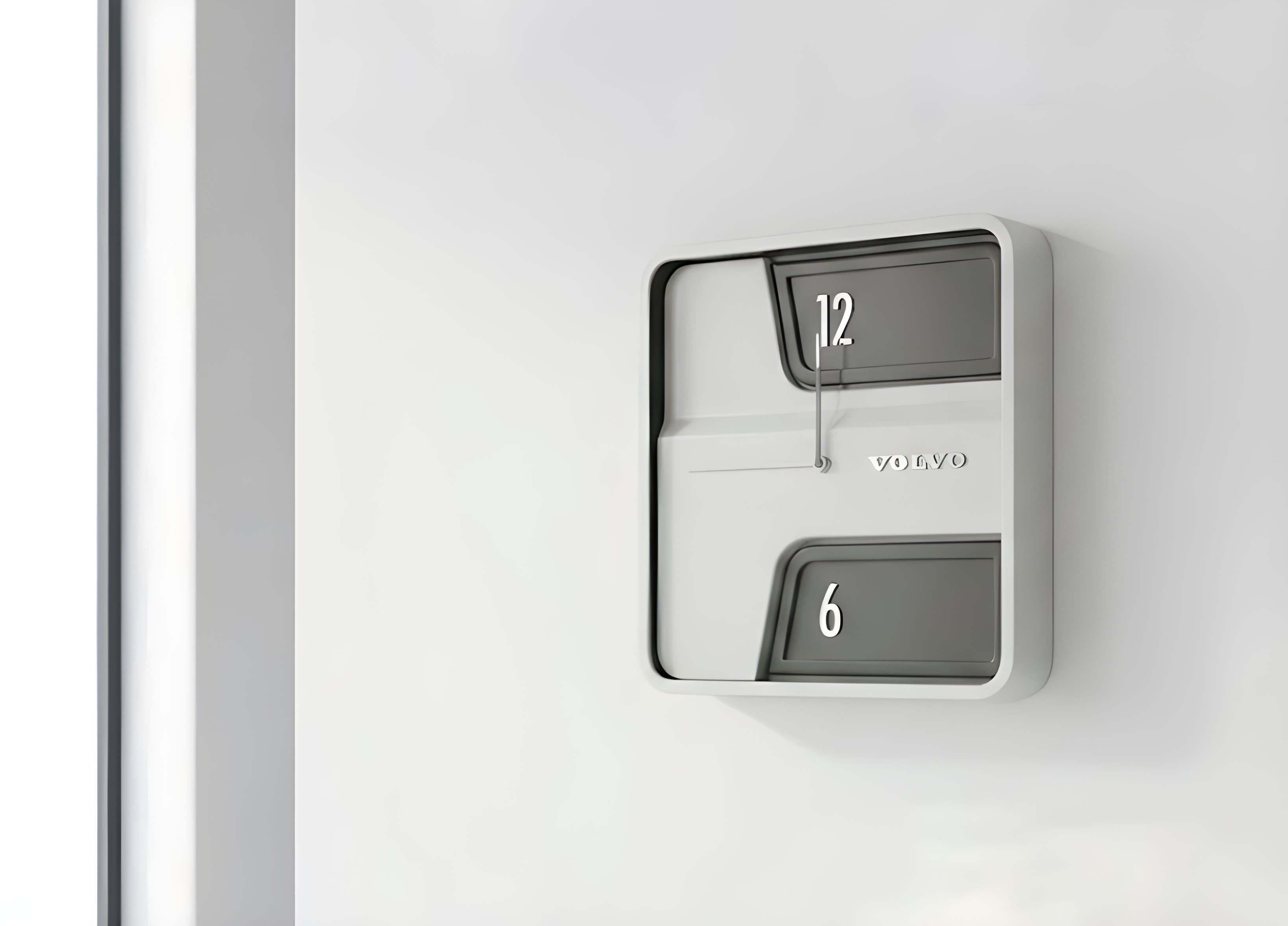 El reloj montado en una pared blanca, visto en ángulo: cuerpo color crema, dos ventanas en relieve que dejan ver el 12 y el 6, y el logotipo de Volvo grabado en bajorrelieve sobre la caja.